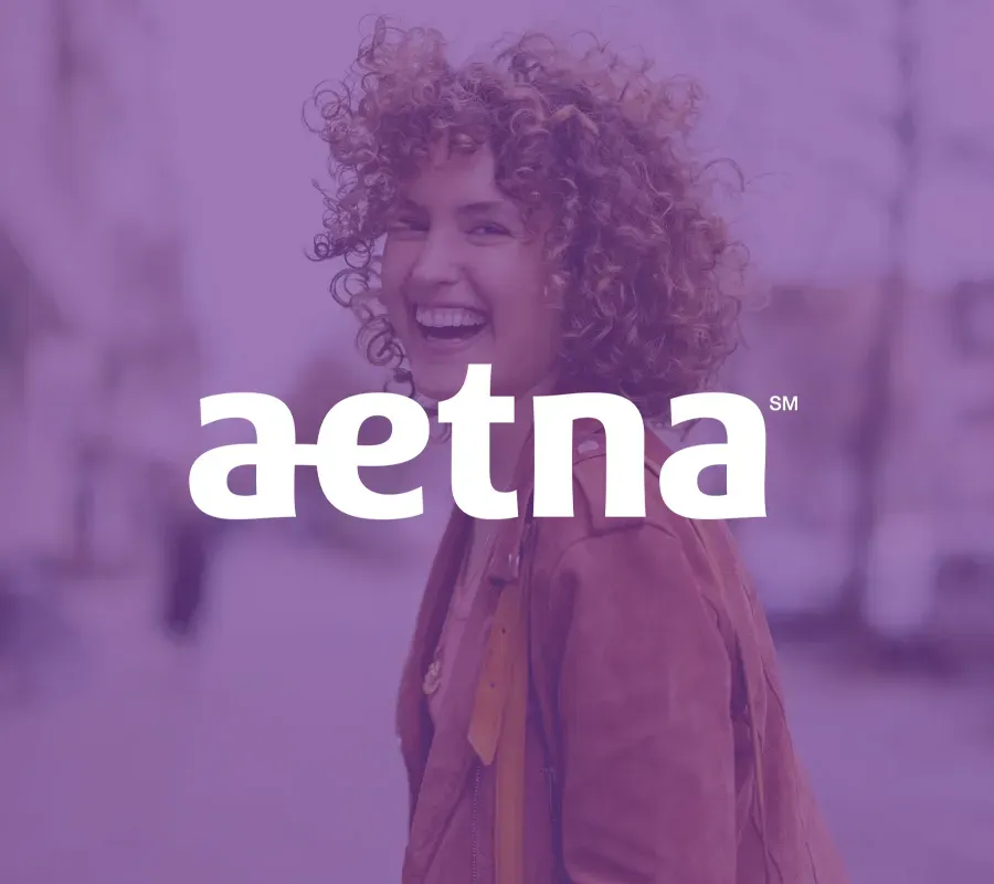 Smiling curly-haired woman on a street with a purple-tinted Aetna logo overlay.