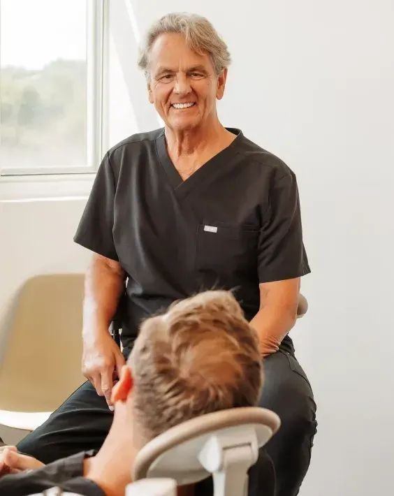 Smiling healthcare provider in dark scrubs sitting with a patient in a chair.