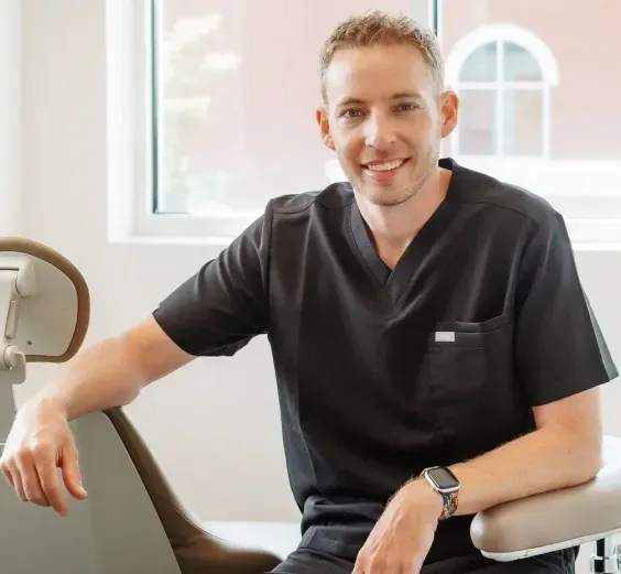 Smiling man in black scrubs sitting on a dental chair indoors.