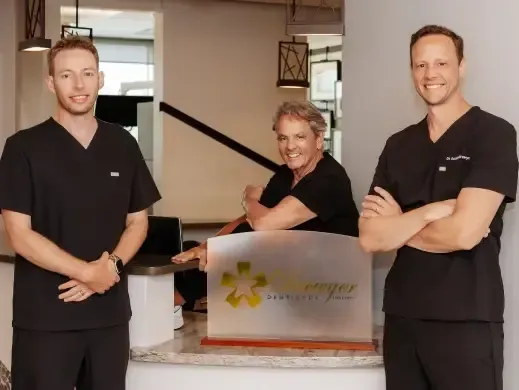 Three men in black scrubs pose behind a dental office reception desk sign.